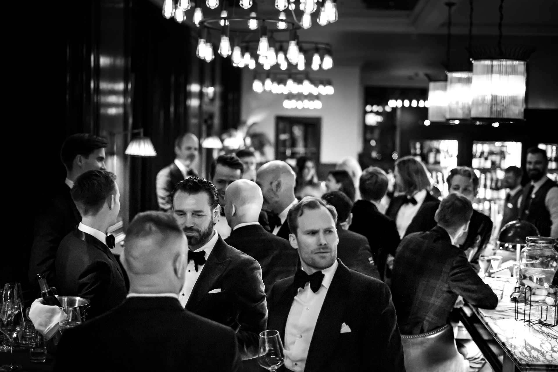 Members in black tie in conversation along a bar under filament chandeliers